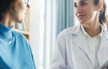 Médica conversa com paciente durante consulta, segurando uma prancheta em um ambiente claro de atendimento em saúde.